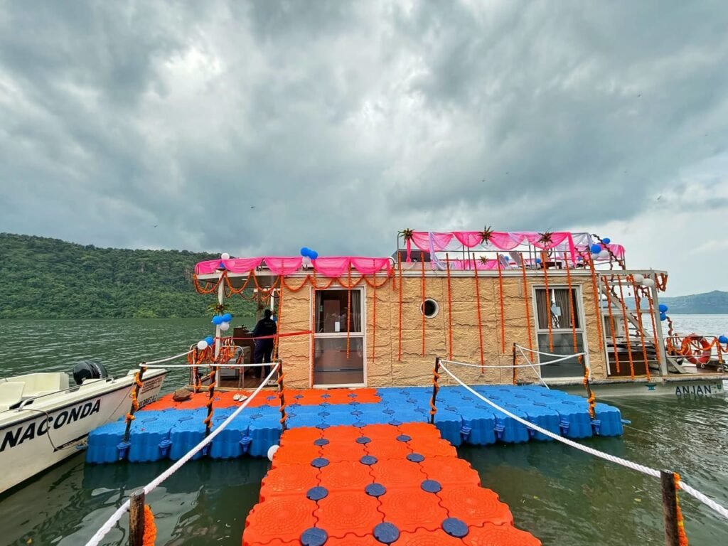 करमचट डैम की झील में तैरती हुई एक लक्ज़री हाउसबोट, पीछे हरी-भरी पहाड़ियां और झरनों का नज़ारा।